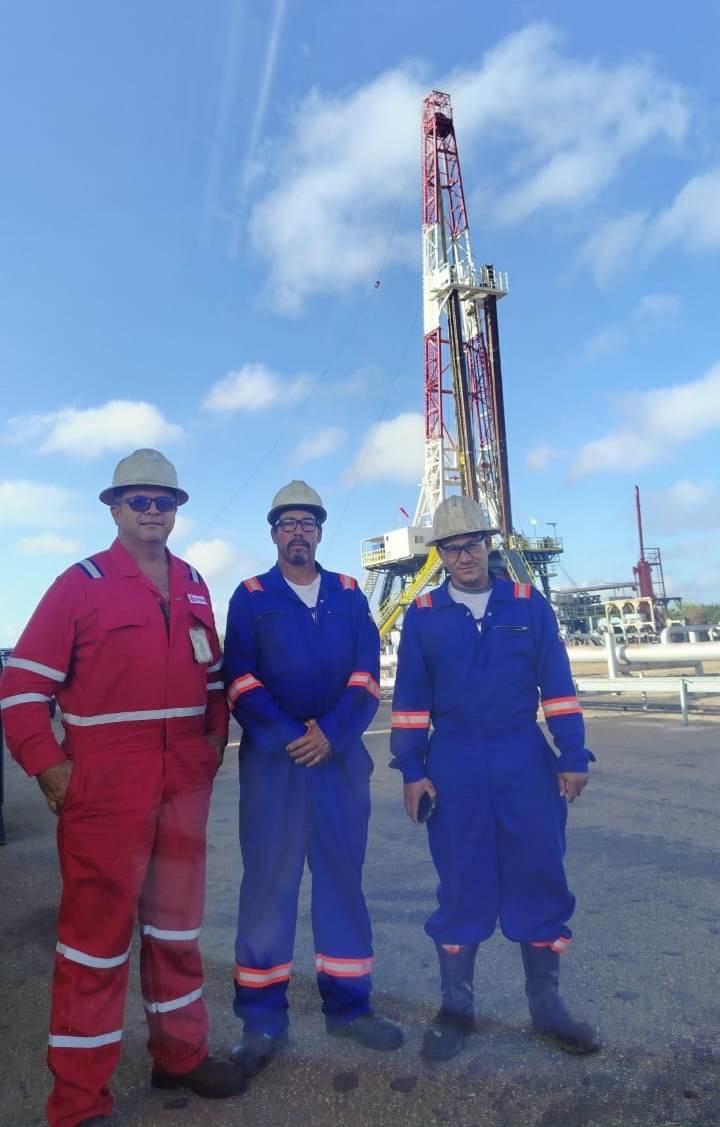 Tres integrantes de Farfar C.A. con braga y casco, con una torre de perforación al fondo
